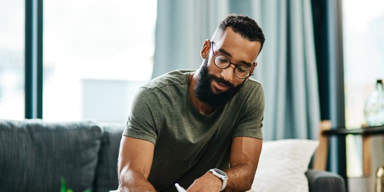 A young muscular man with a beard and glasses, sits on his couch completing his will.